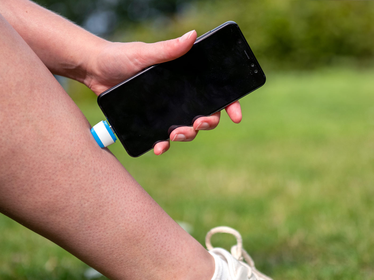 Eine Person hält im Freien auf einer Wiese ein Smartphone an ihr Bein, an dem das kleine bite away® pro Gerät befestigt ist.